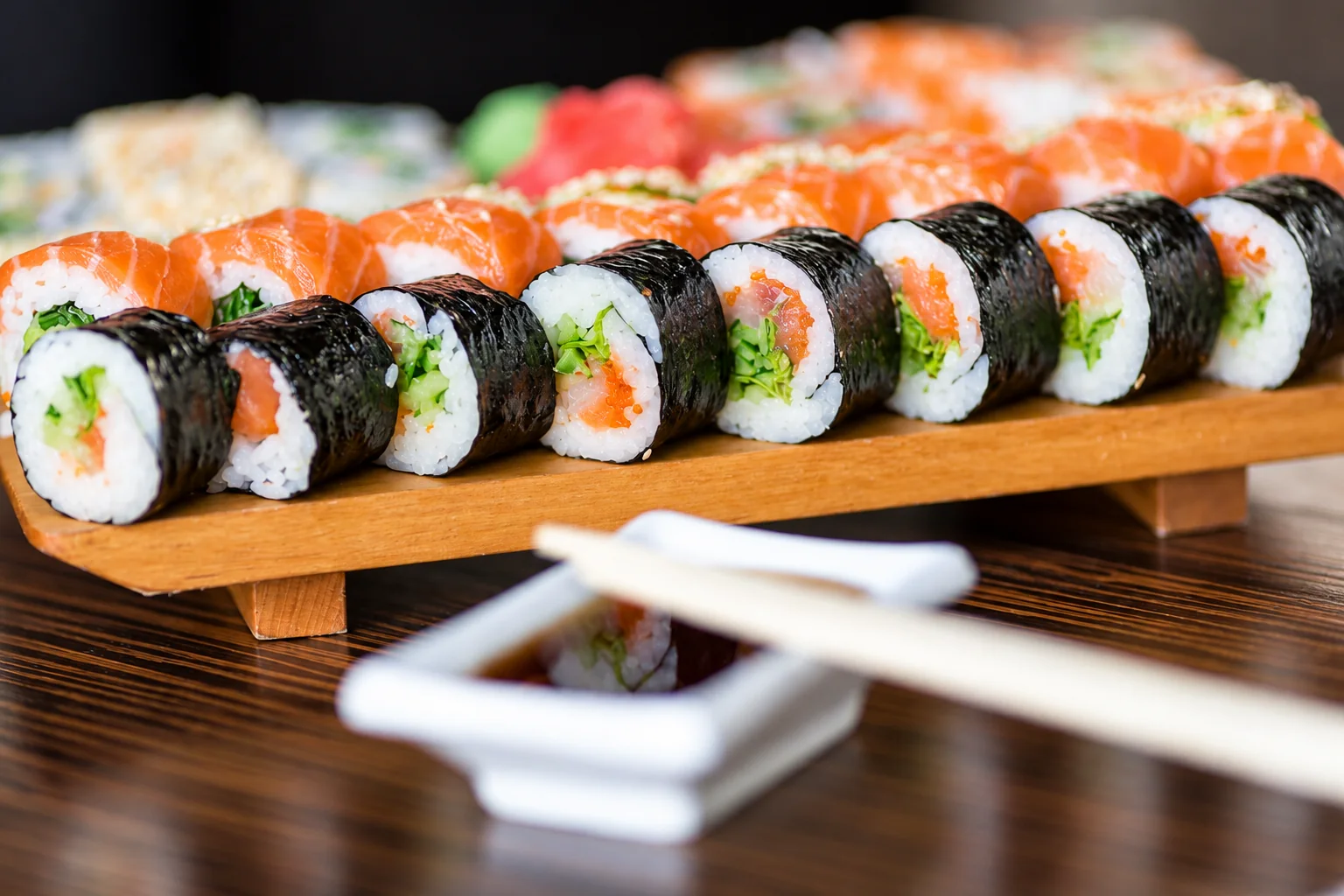 Maki lined up on a wooden stand, sauce dish and chopsticks in the foreground.