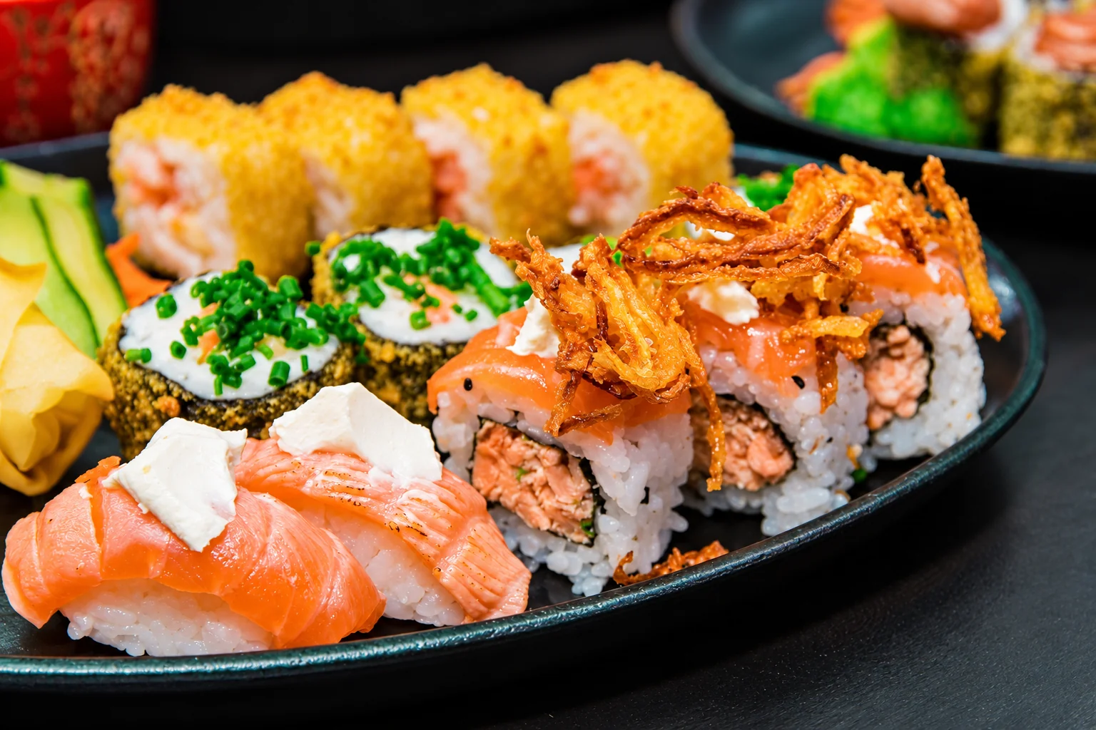 Salmon sushi, maki and rolls topped with fried onions on a black plate.
